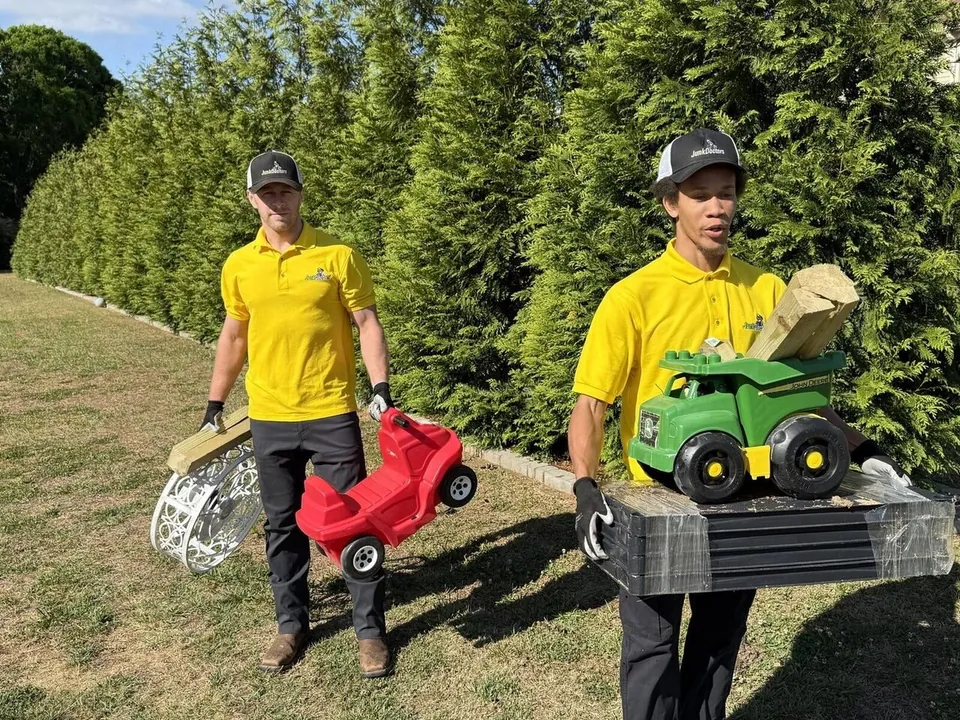 Two Junk Doctors crew members carrying old toys and a grill across a yard to the truck